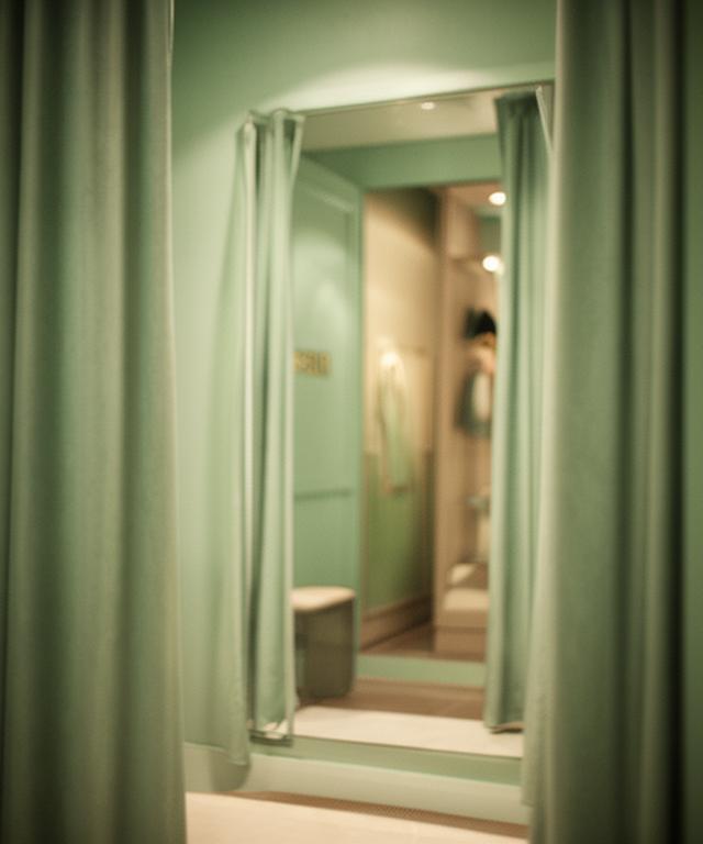 A quiet eveningwear boutique fitting room with a curtain, mirror, and soft green glass reflections